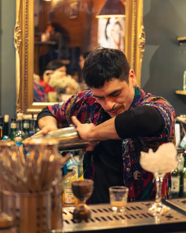 Bartender de camisa estampada colando un trago en la barra, cobertura de noche en un bar de Valparaíso