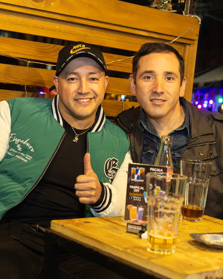 Dos hombres sonríen junto a sus tragos, uno con el pulgar arriba, en un bar de Valparaíso, cobertura de noche