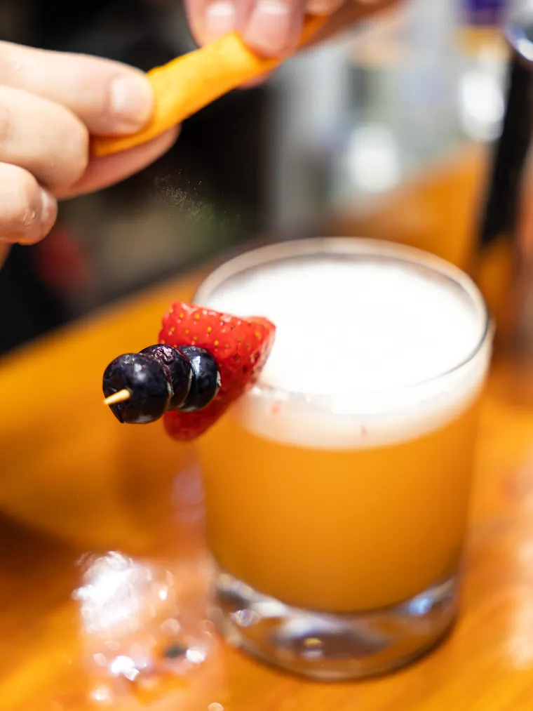 Cóctel espumoso con brocheta de frutilla y arándanos mientras se exprime una cáscara de naranja encima