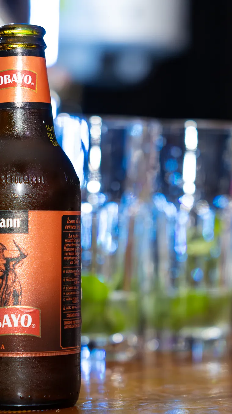 Botella de cerveza sobre la barra y vasos desenfocados al fondo, fotografía gastronómica en Valparaíso