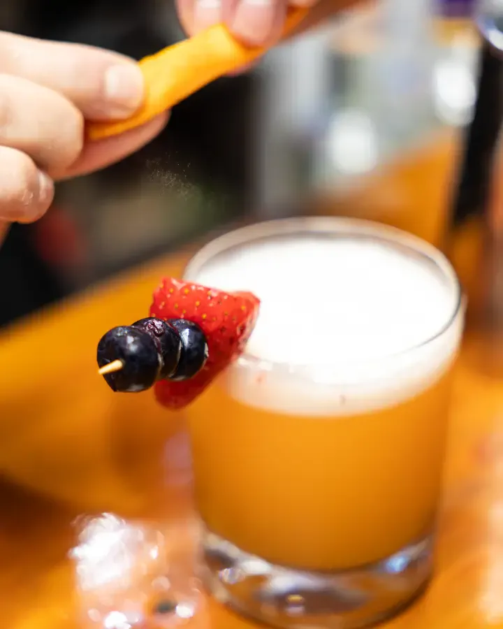 Trago naranja con espuma, brocheta de frutilla y arándanos y una mano espolvoreando encima, fotografía de producto