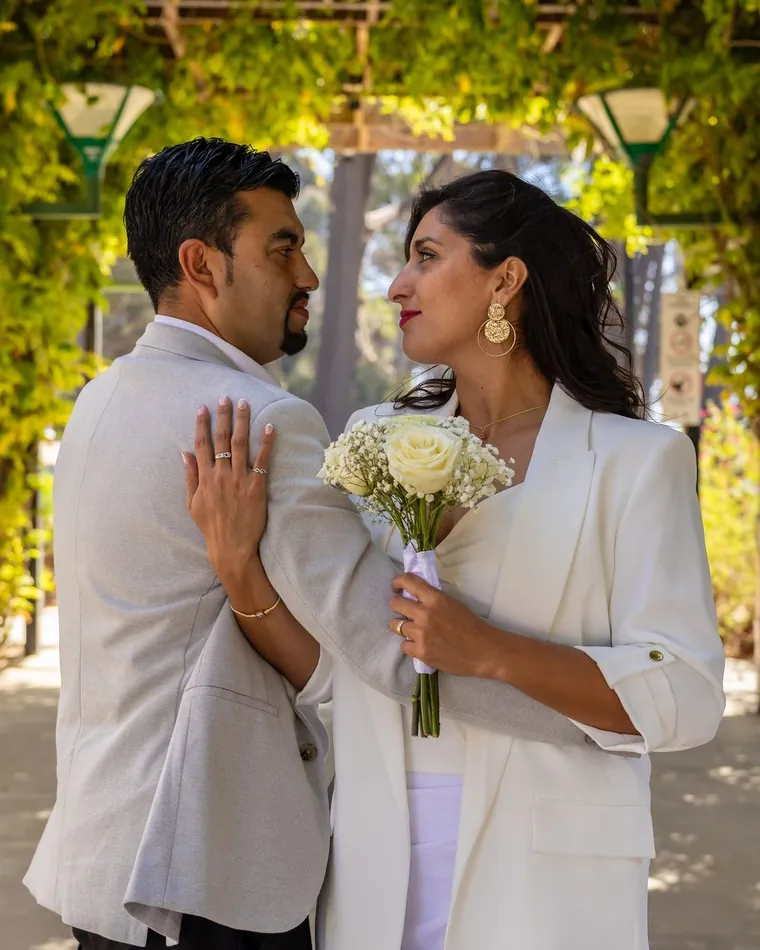 Novios mirándose bajo una pérgola verde, ella de blanco con ramo de rosas, fotografía de matrimonios