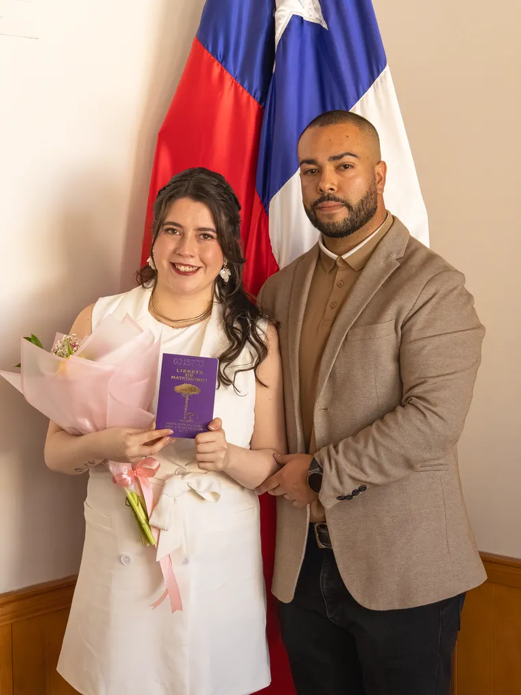 Los novios posan con la libreta de matrimonio y el ramo frente a la bandera de Chile