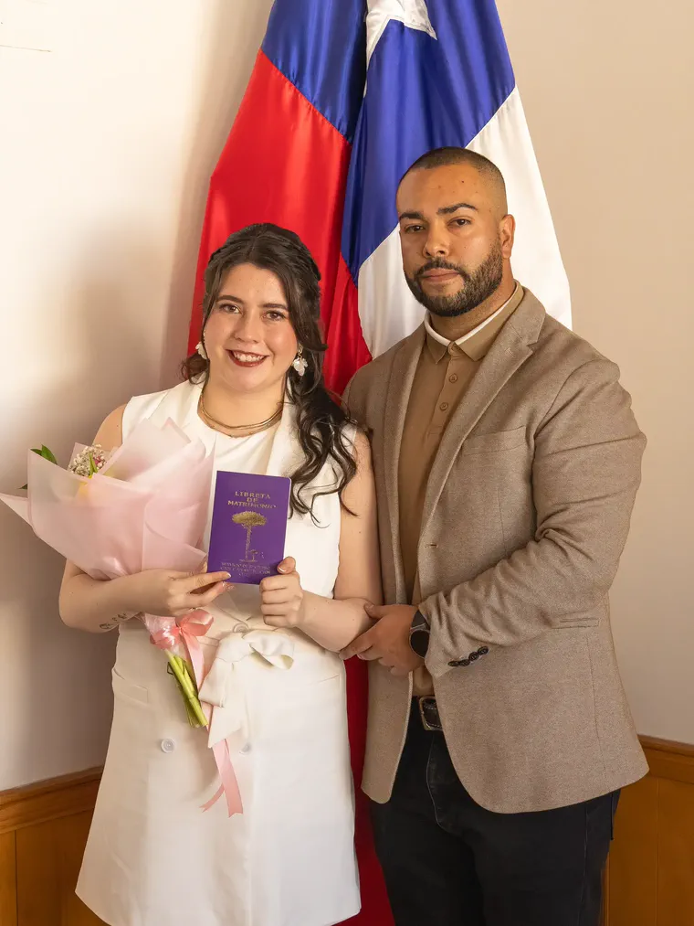 Los novios posan con la libreta de matrimonio y el ramo frente a la bandera de Chile