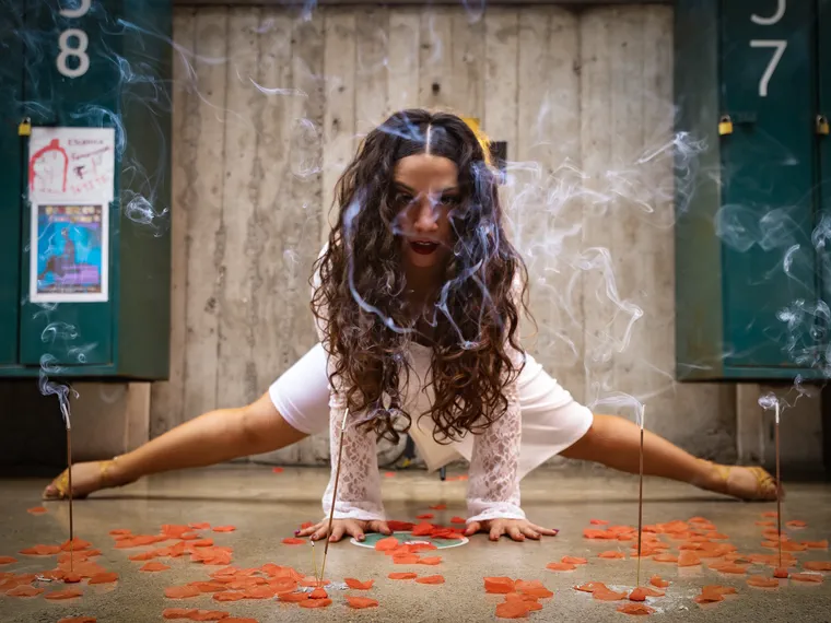 Artista de vestido blanco agachada en pose de baile entre humo de incienso y pétalos rojos, sesión en locación