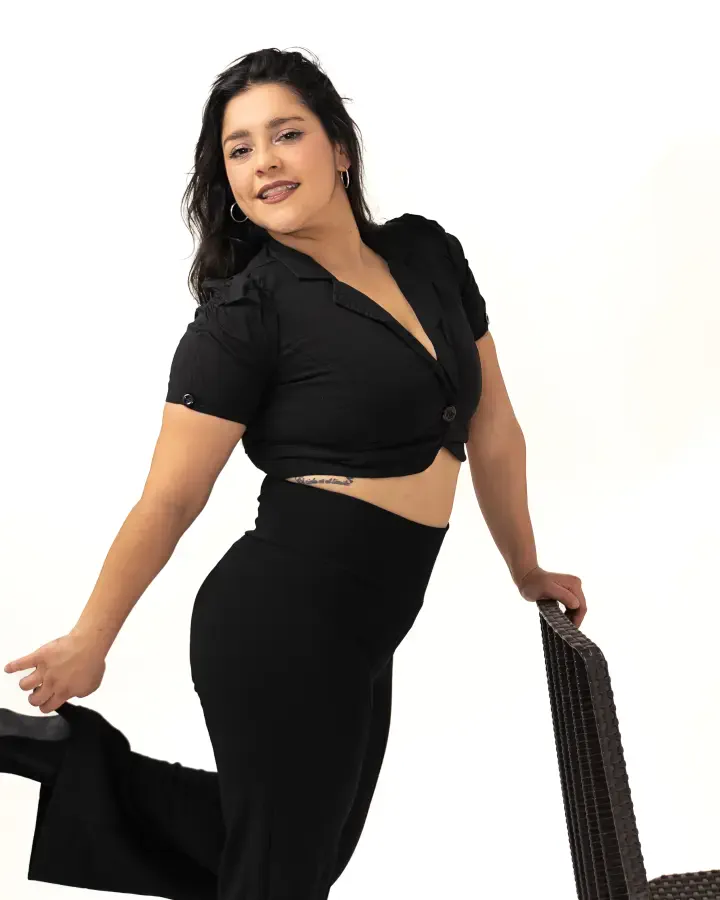 Mujer sonríe apoyada en una silla de mimbre, de top y pantalón negros, en sesión de retrato sobre fondo blanco